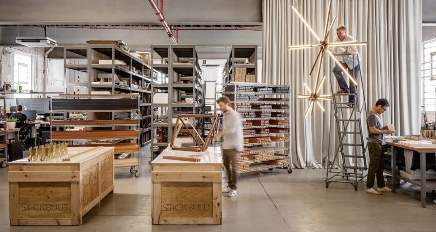 Workers assembling wooden light fixtures in a spacious, organized workshop with shelves and materials around.