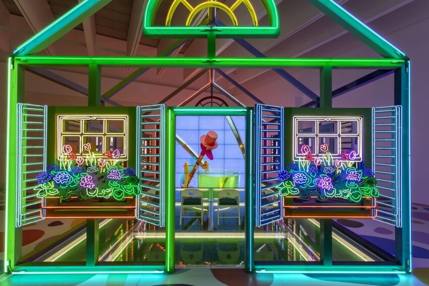 Bright neon art installation of a colorful house with open windows and flowers, featuring a character in the background.