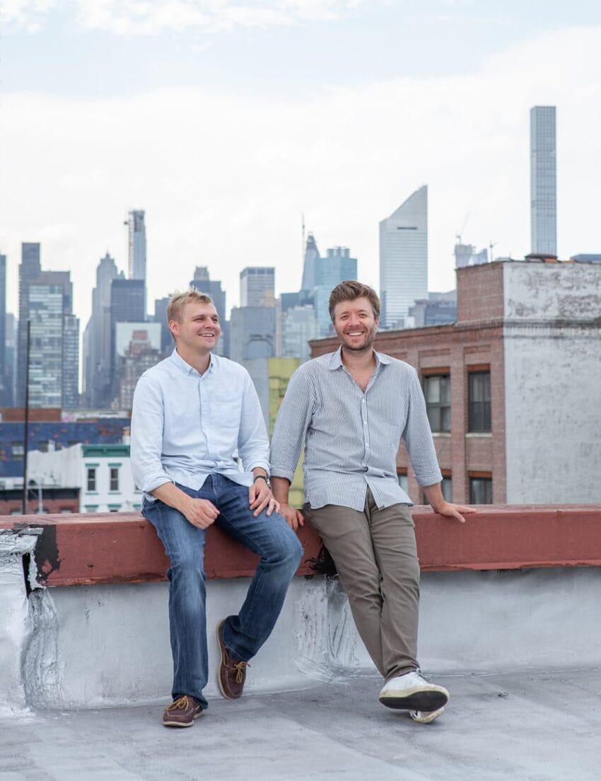 Two people sitting on a rooftop edge with a city skyline in the background, one wearing jeans and the other in khakis.