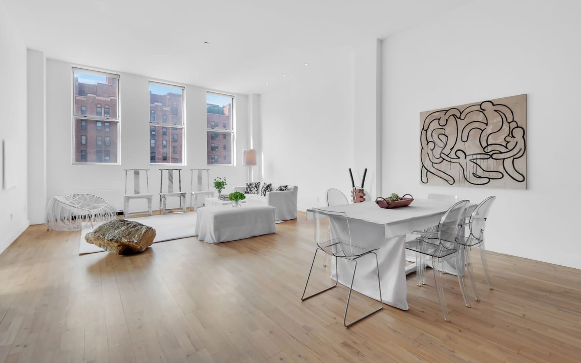 Spacious modern living room with large windows, white furniture, abstract wall art, and a dining table with transparent chairs.