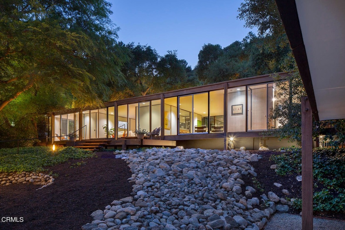 Modern glass house with illuminated interior and surrounding trees at dusk