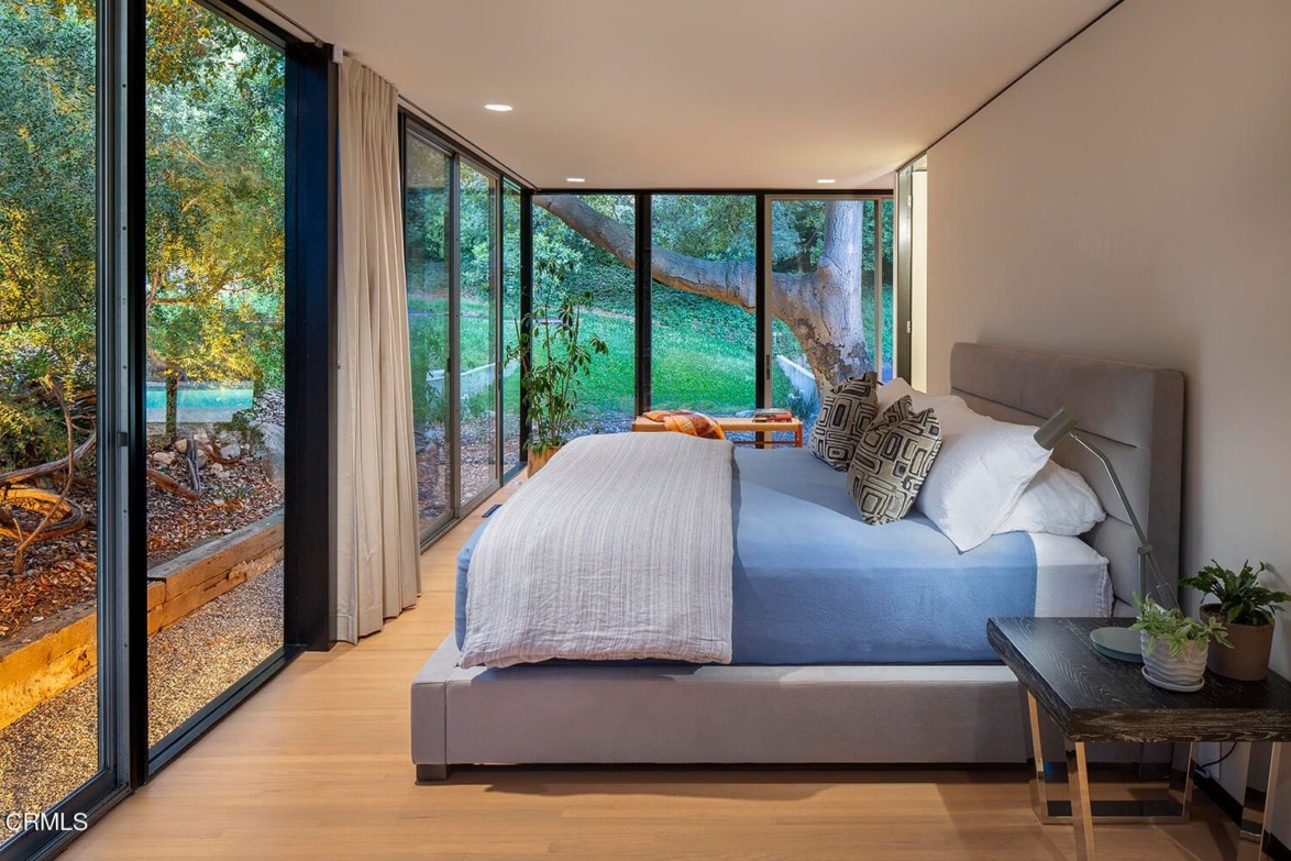 Modern bedroom with large windows, nature view, blue bedding, and stylish decor. Light wood flooring and small plants add warmth.