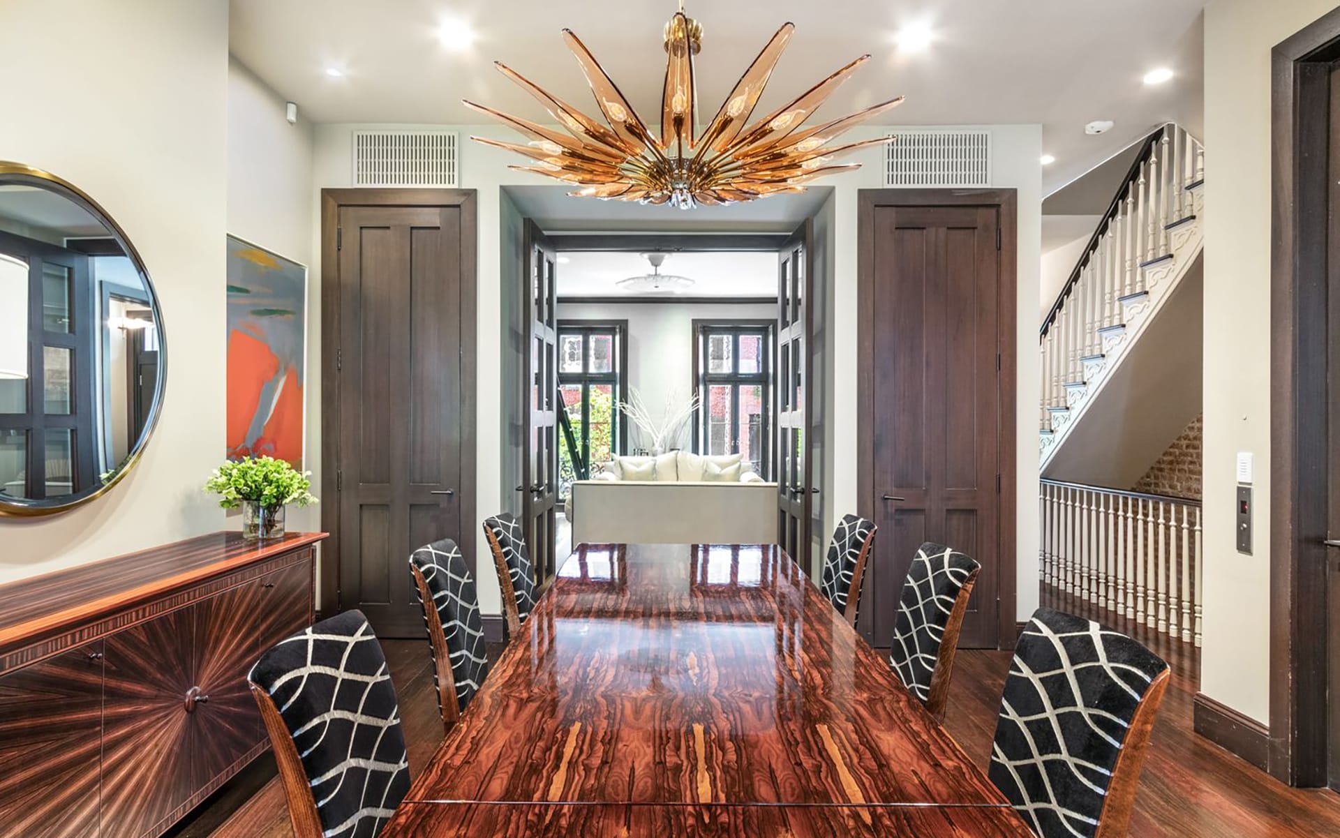 Dining room with elegant wooden table, patterned chairs, modern chandelier, and view into living room and staircase.