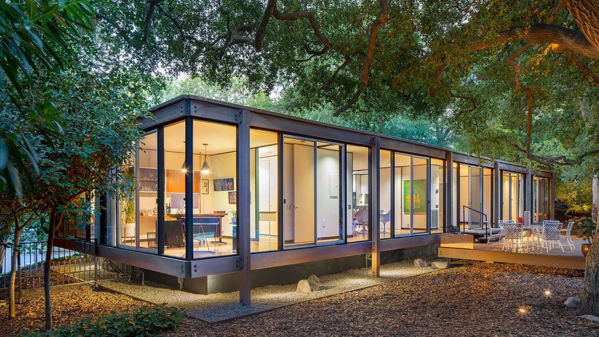 Modern glass-walled house surrounded by trees with a spacious wooden deck adorned with outdoor furniture.