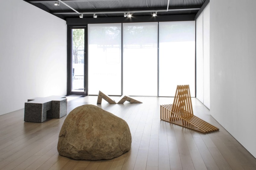 Minimalist art gallery with natural light, featuring wooden and stone sculptures on a light wood floor.