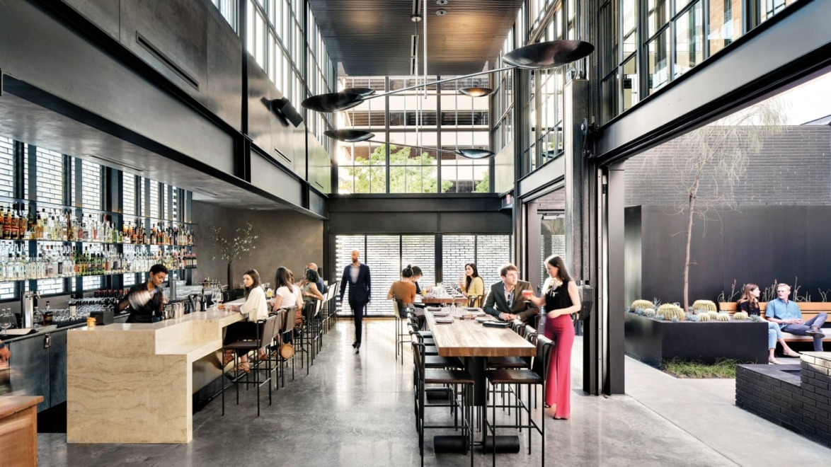 Modern restaurant interior with people dining at tables and bar counter, large windows, and an outdoor seating area.