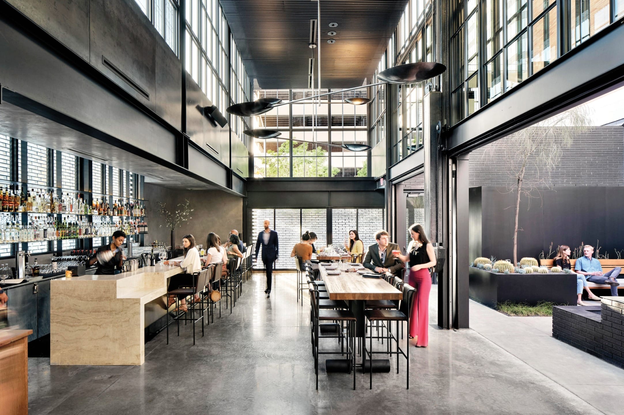 Modern restaurant interior with people dining at tables and bar counter, large windows, and an outdoor seating area.