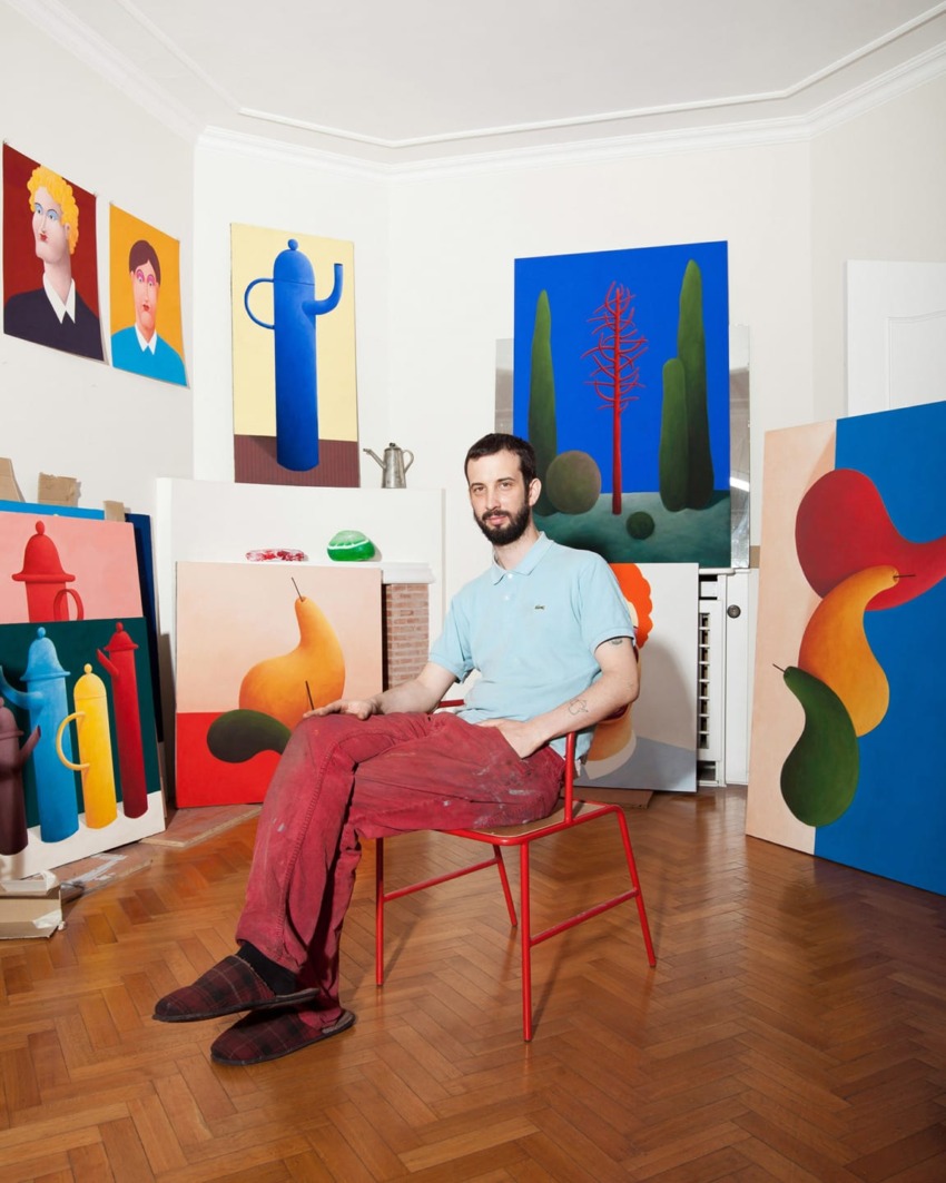 Artist sitting in a studio surrounded by colorful abstract paintings and portraits on walls and floor.