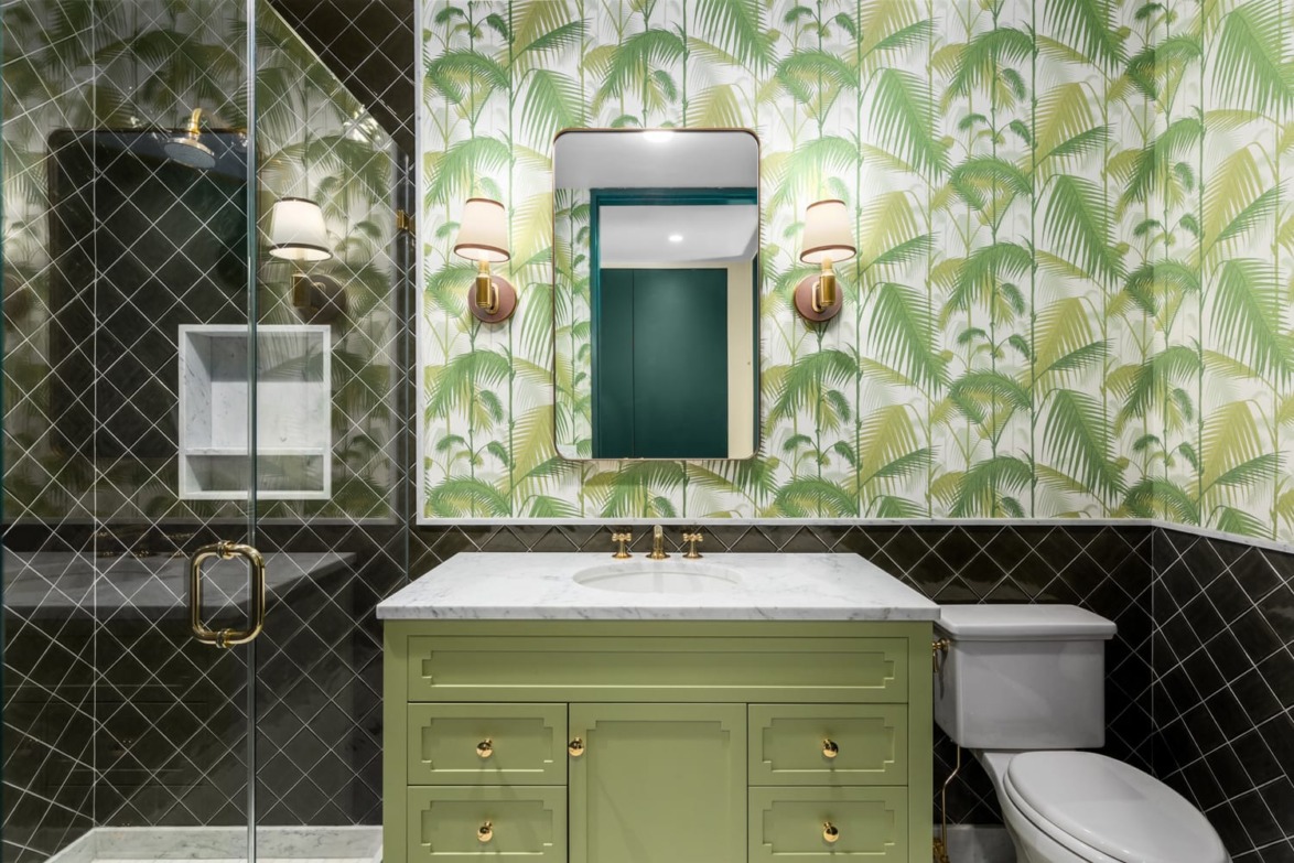 Bathroom with tropical leaf wallpaper, green vanity, marble countertop, glass shower, and modern lighting fixtures.