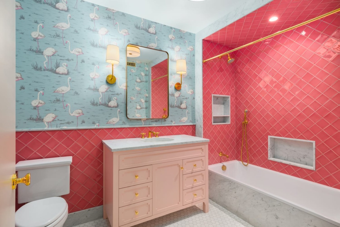 Colorful bathroom with flamingo wallpaper, pink vanity, gold fixtures, pink tile bathtub, and white toilet.