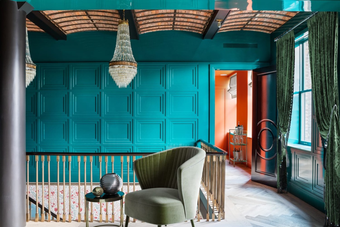 Elegant interior with a green armchair, teal paneled walls, chandeliers, and a wood ceiling, bathed in natural light.