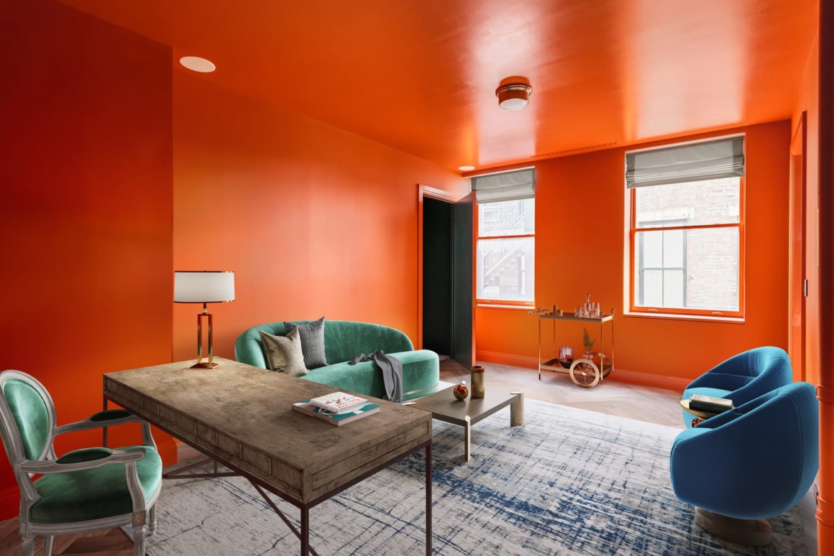 Modern orange-walled room with green couch, blue chairs, wooden desk, and decor accents near large windows.