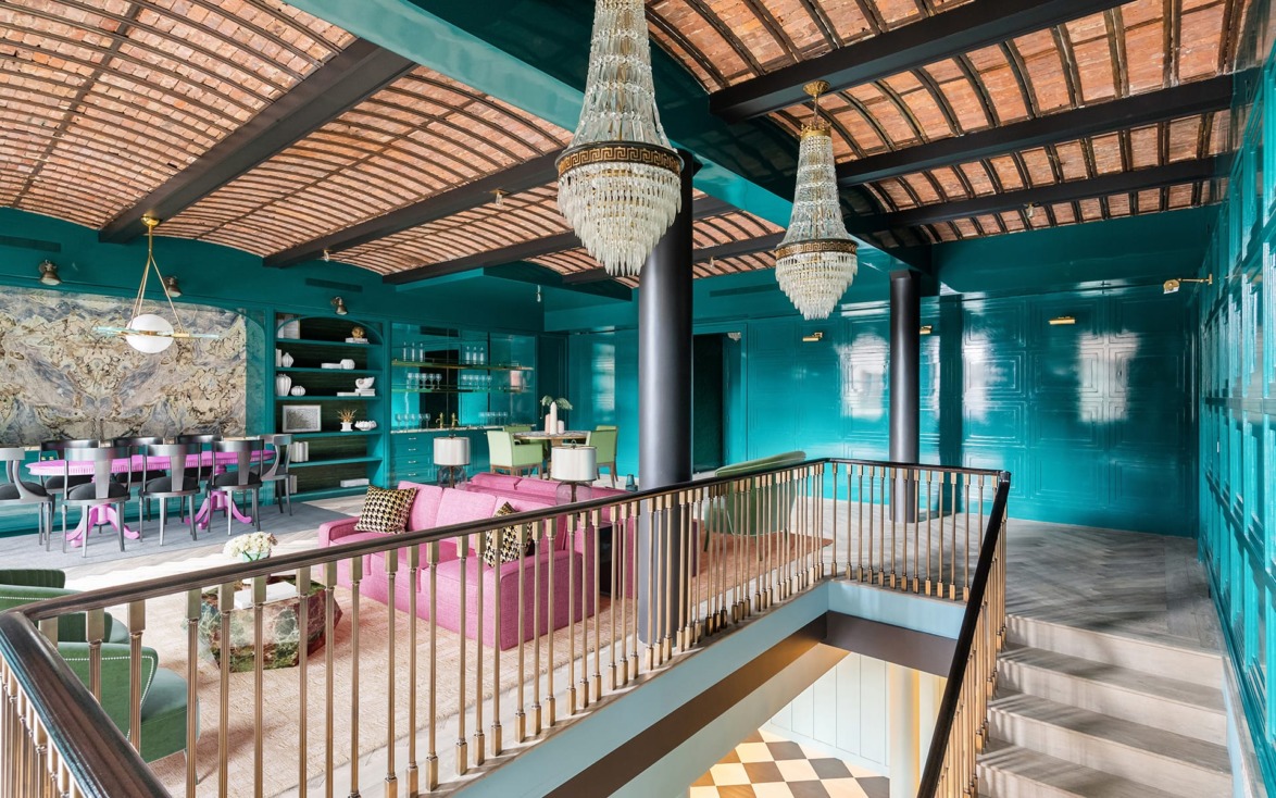 Luxurious modern loft with teal walls, exposed brick ceiling, elegant chandeliers, and eclectic pink and green furniture.