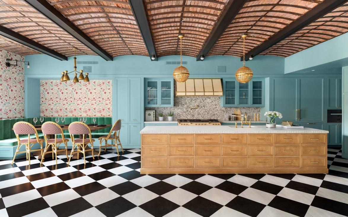 Modern kitchen with checkered floor, light blue cabinets, floral wallpaper, wooden island, gold accents, and green seating nook.
