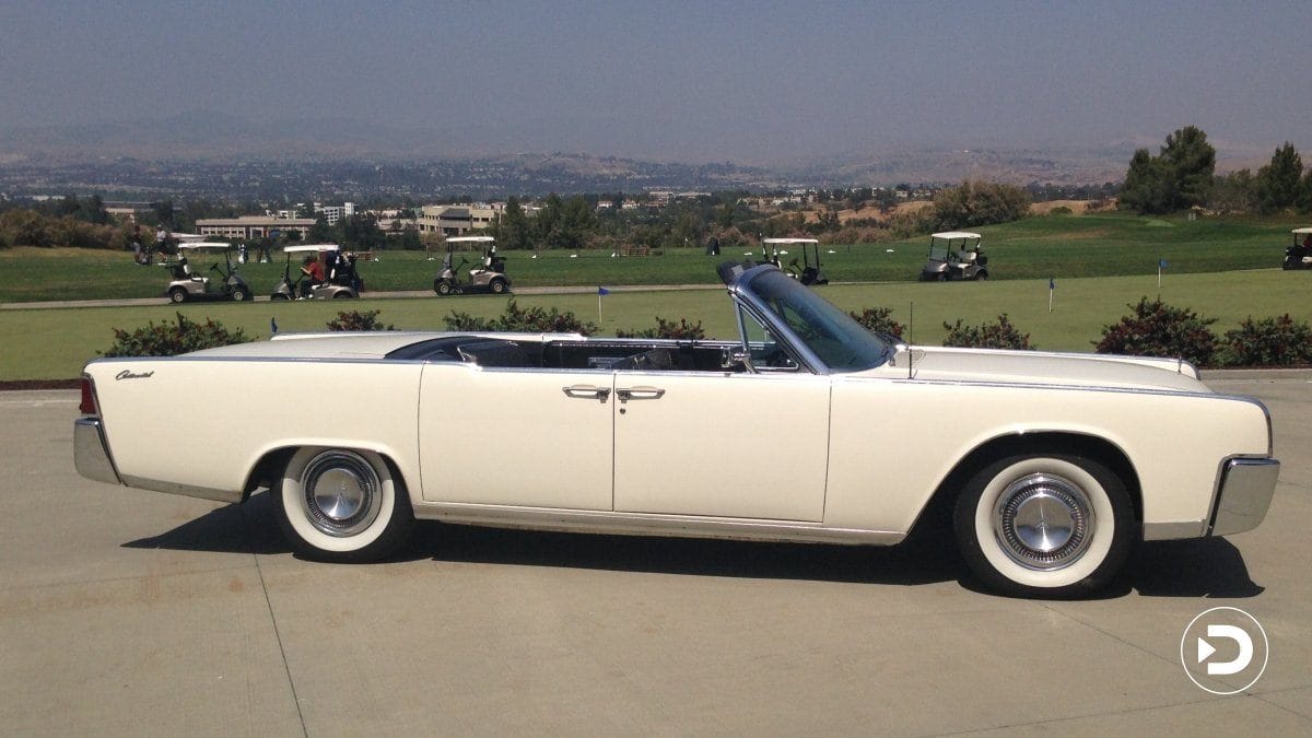 White vintage convertible car parked on concrete surface with a golf course and hills in the background.