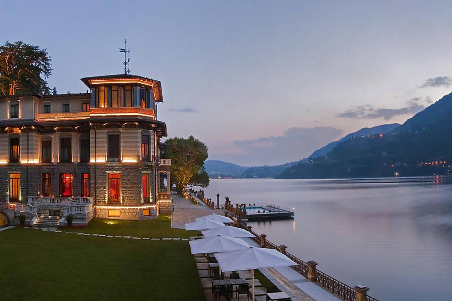 Lakeside villa at dusk with lit windows, surrounded by mountains and a calm lake, featuring an elegant outdoor terrace.