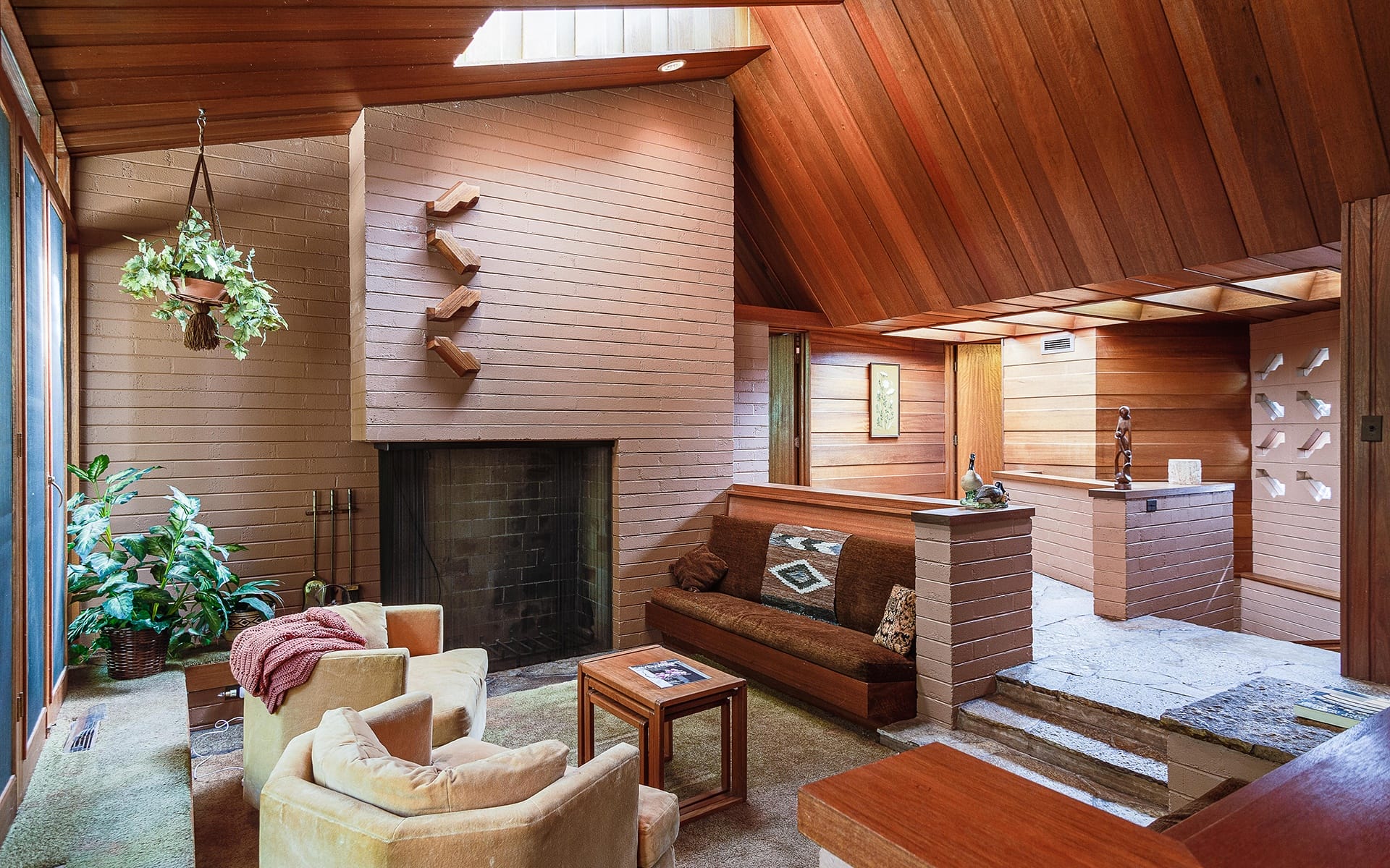 Mid-century modern living room with wooden ceilings, fireplace, armchairs, and plants.