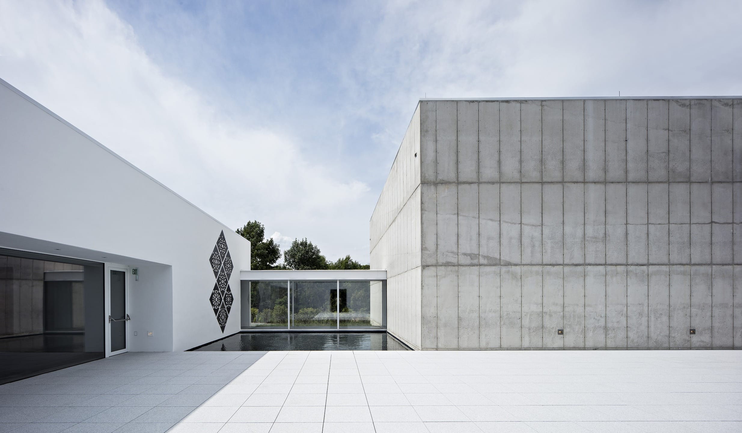Modern minimalist building with concrete and glass design, reflecting pool, and open courtyard under a cloudy sky.