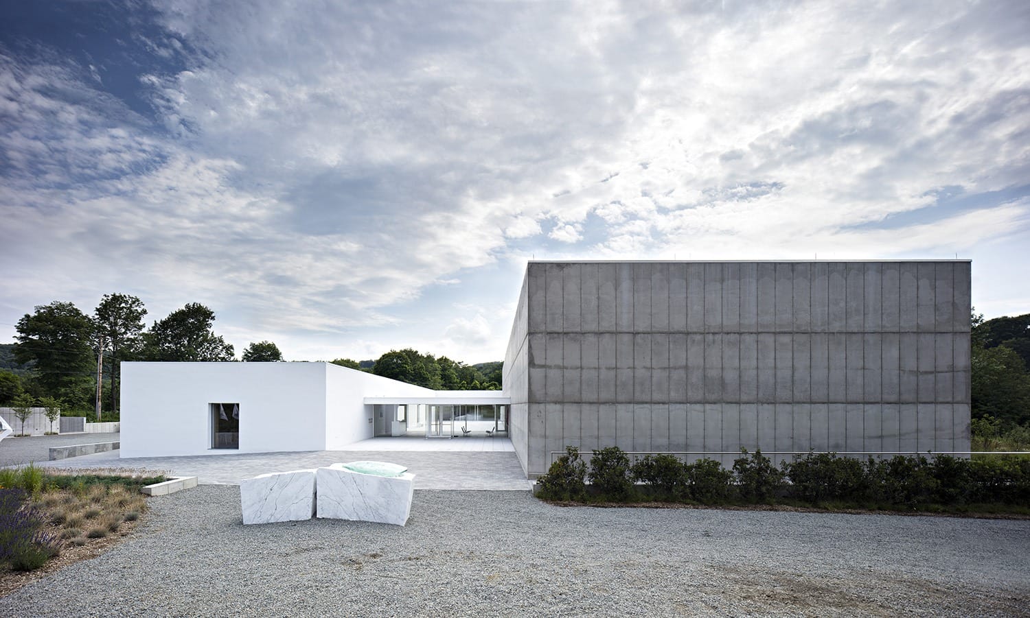 Modern minimalist building with concrete and white structures, surrounded by gravel and greenery under a cloudy sky.