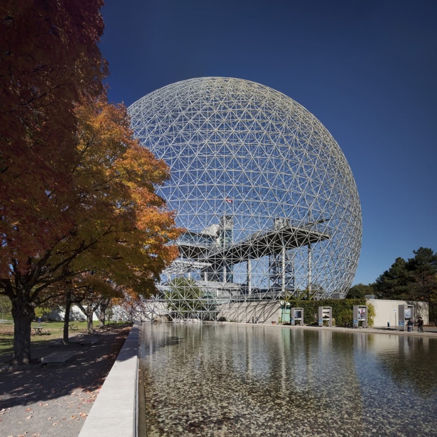Geodesic dome structure with a reflective pond and autumn trees under a clear blue sky.