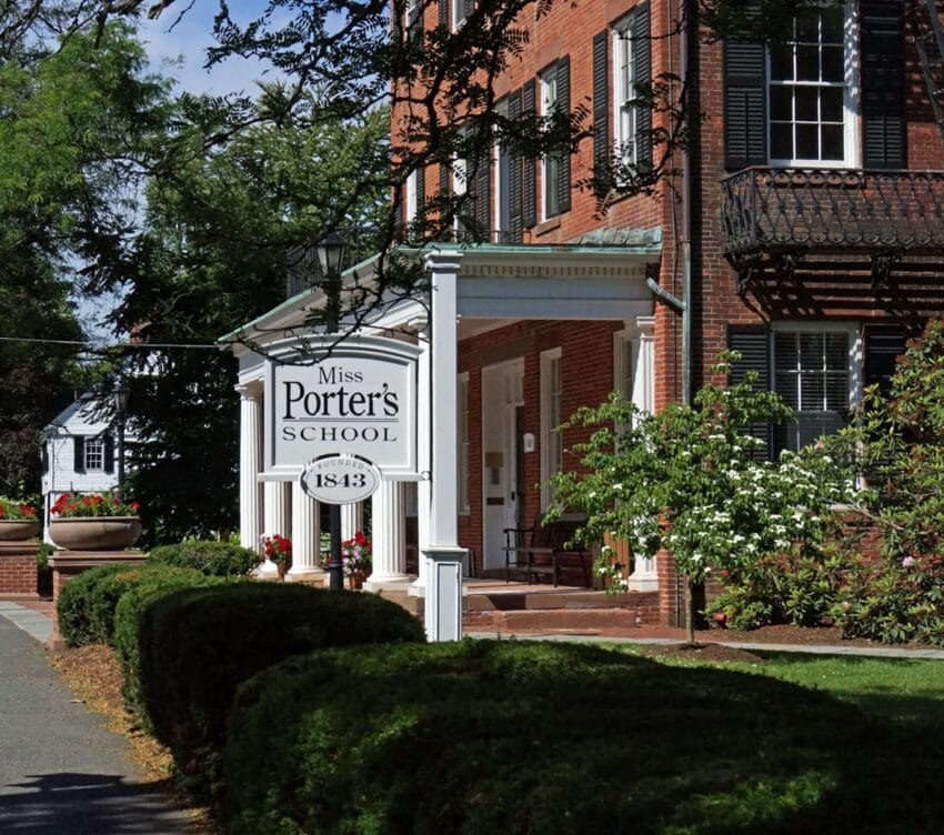 Historic brick building with a sign reading "Miss Porter's School 1843" surrounded by greenery and a blue sky.