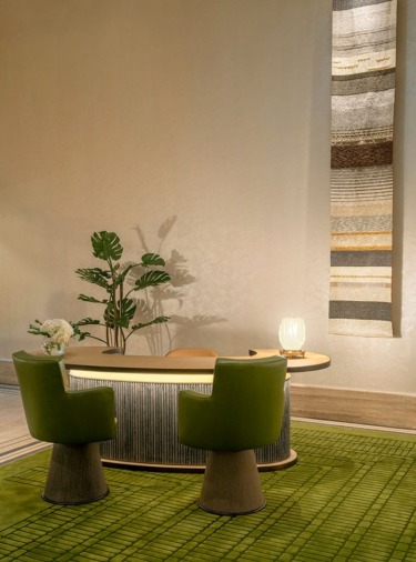 Elegant lobby with a curved desk, two green chairs, a tall plant, and a textured wall hanging on a green rug backdrop.