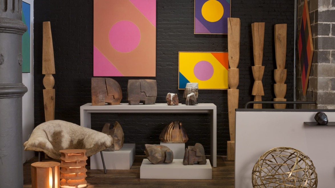 Gallery room with abstract paintings, wooden sculptures, and modern decor elements displayed on a table and floor.