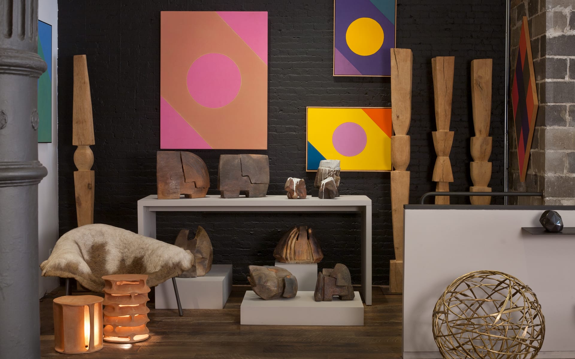 Gallery room with abstract paintings, wooden sculptures, and modern decor elements displayed on a table and floor.