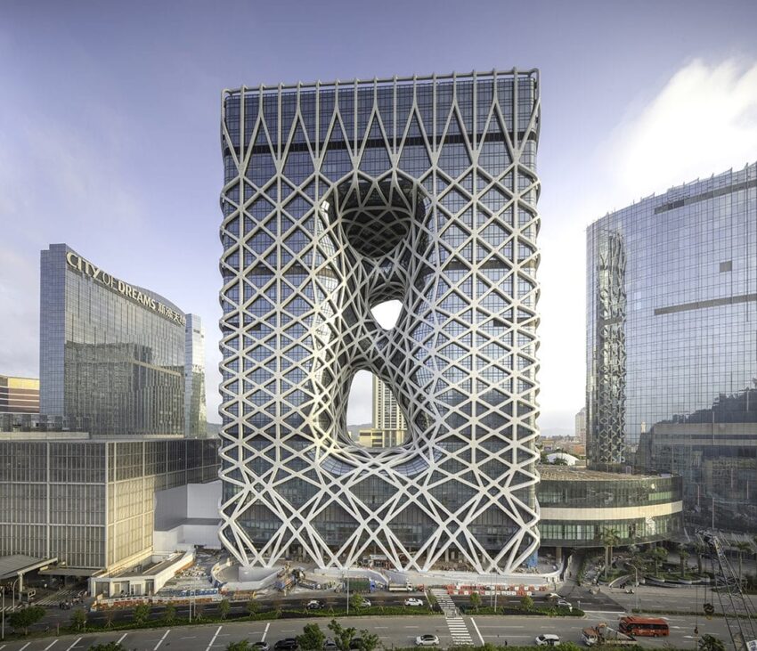Modern building with intricate geometric facade and central void, surrounded by other city skyscrapers against a clear sky.