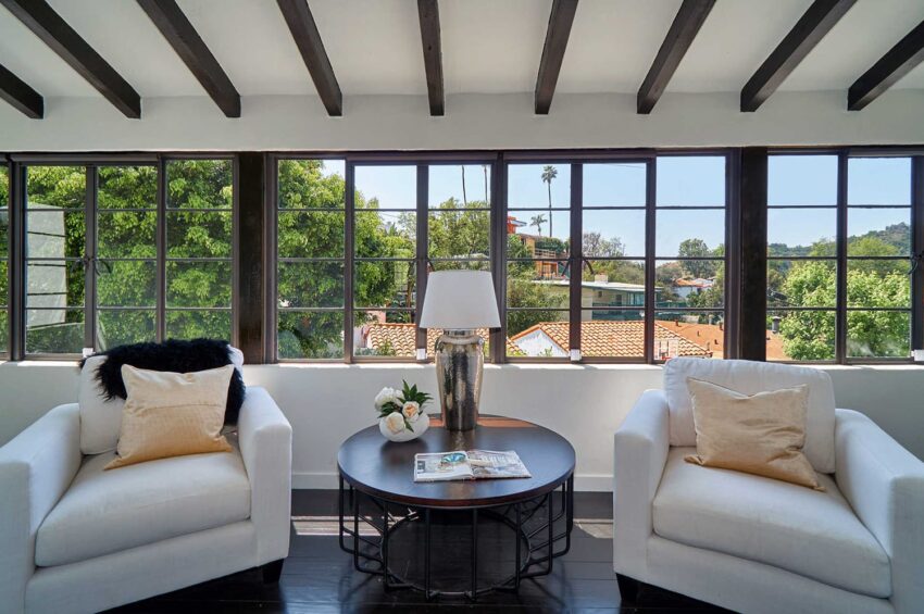 Cozy sitting area with two white armchairs, a round table, decorative pillows, and a window view of green trees.
