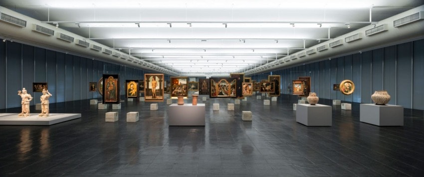 Art gallery interior with various sculptures and paintings displayed on stands and walls. Bright lighting and wide open space.