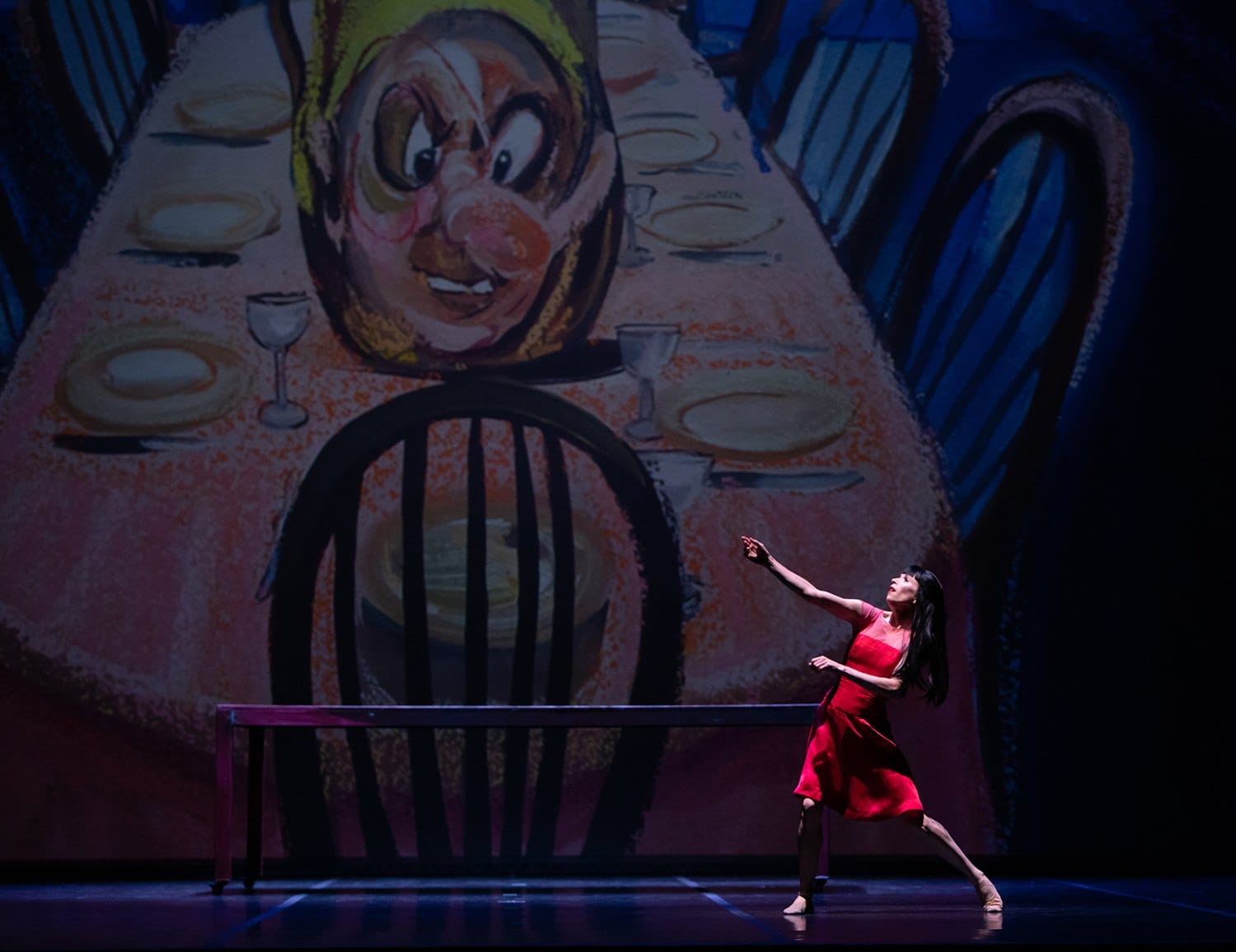 Dancer in red dress performs in front of animated backdrop with large cartoonish face and dining table setting.