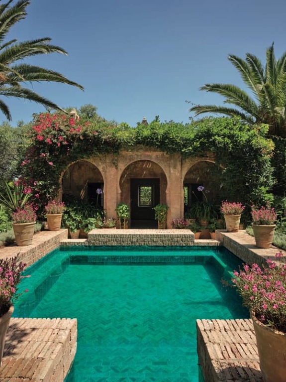 Mediterranean-style garden with turquoise pool, surrounded by lush greenery and terracotta pots under a clear blue sky.