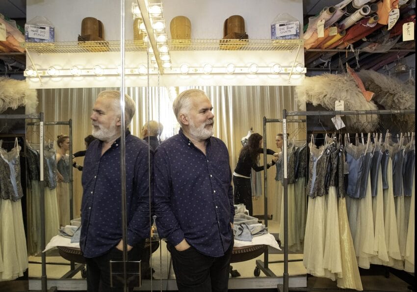 Man standing in a costume shop with mirrors, racks of clothing, and fabric rolls visible.