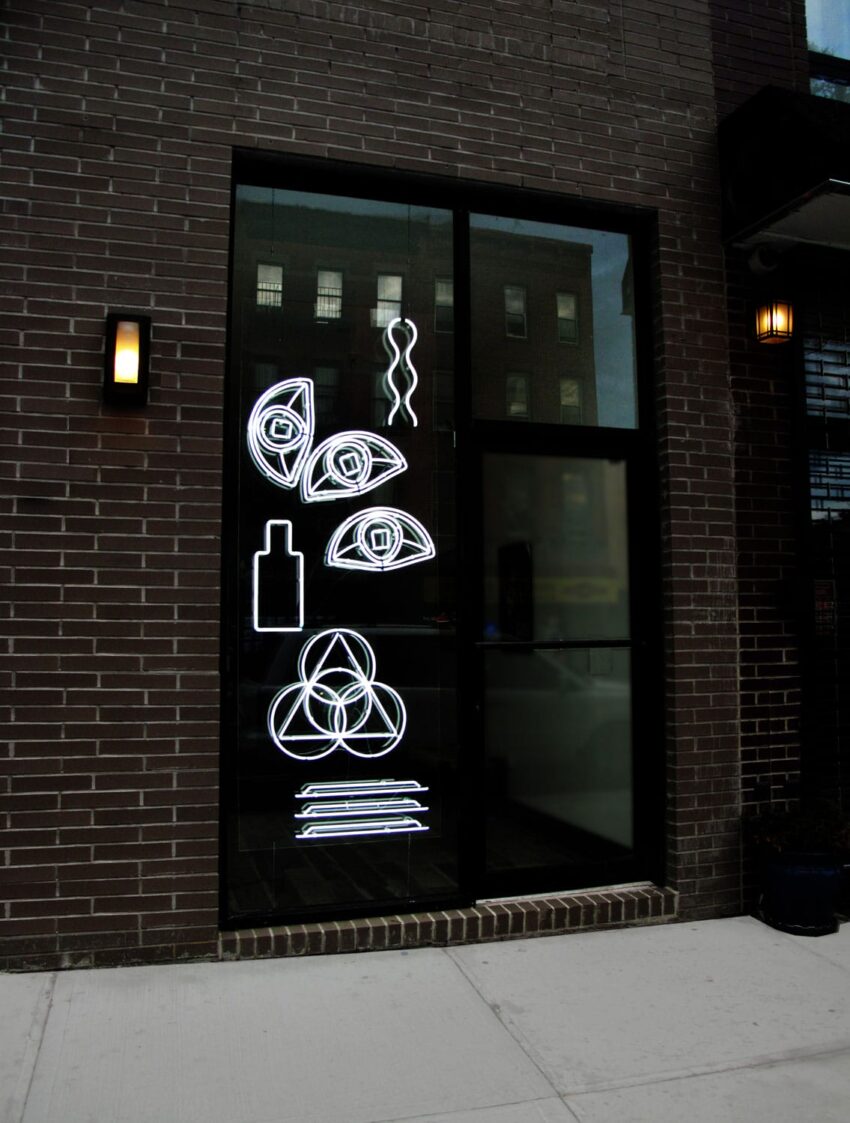 Neon abstract shapes including eyes, squiggles, and a triangle on a window of a dark brick building.
