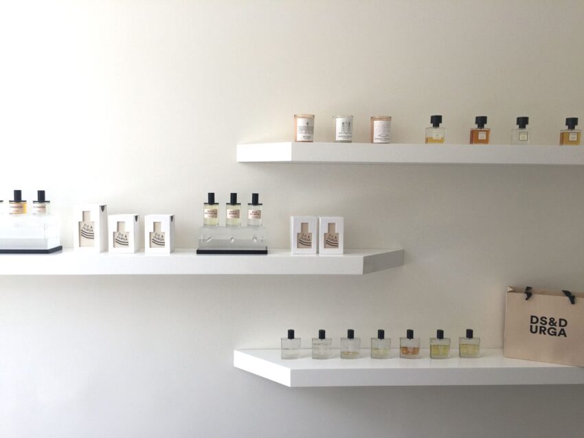 Perfume display on white shelves with various bottles and a branded shopping bag on a minimalist wall background.
