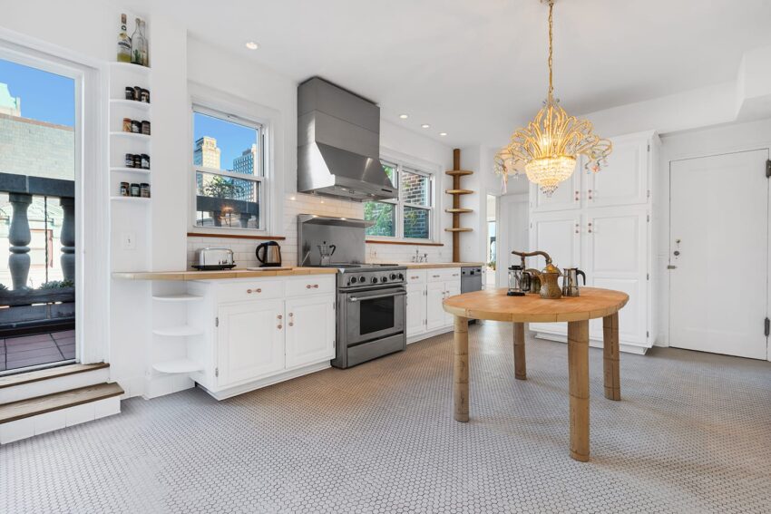 Spacious white kitchen with stainless steel appliances, wooden table, chandelier, and large windows opening to a balcony.