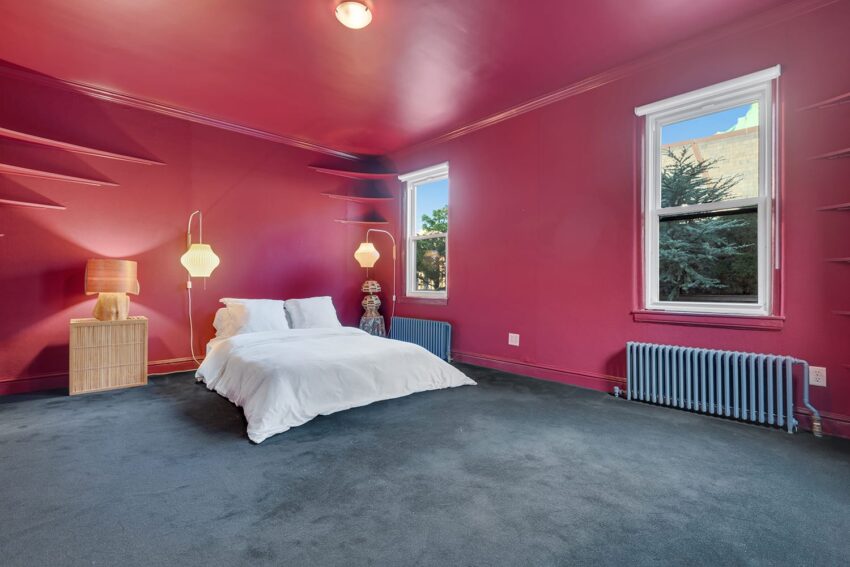 Bedroom with vibrant pink walls, two windows, a white bed, a bedside lamp, and built-in shelves.