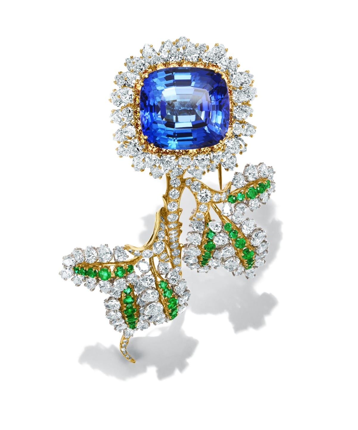 Sapphire brooch with diamond halo and gold leaves, accented by emeralds and diamonds, on a white background.