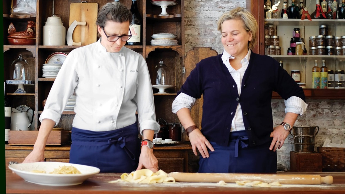 Two chefs wearing aprons are preparing pasta dough on a wooden counter in a rustic kitchen setting with shelves of jars.