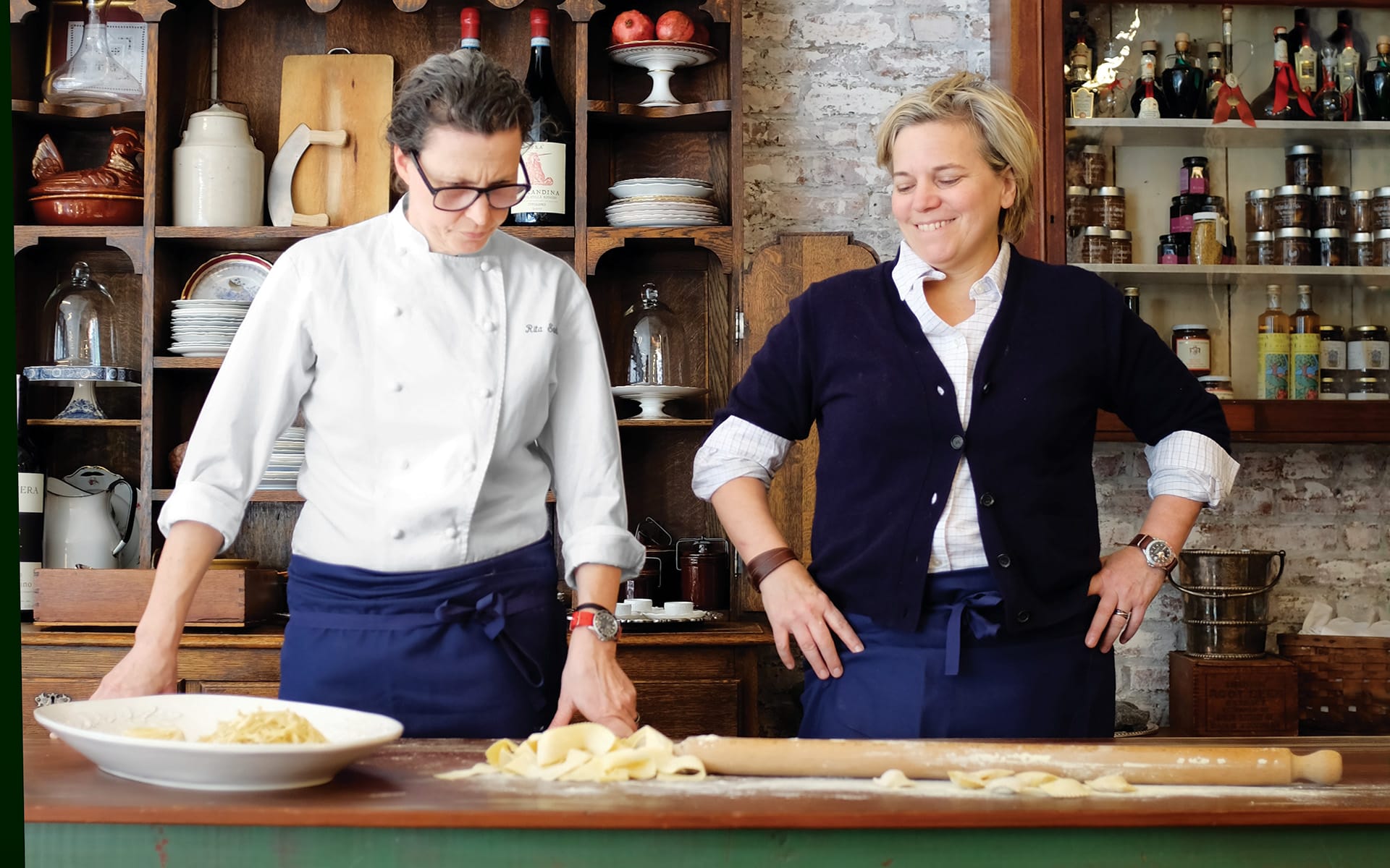 Two chefs wearing aprons are preparing pasta dough on a wooden counter in a rustic kitchen setting with shelves of jars.
