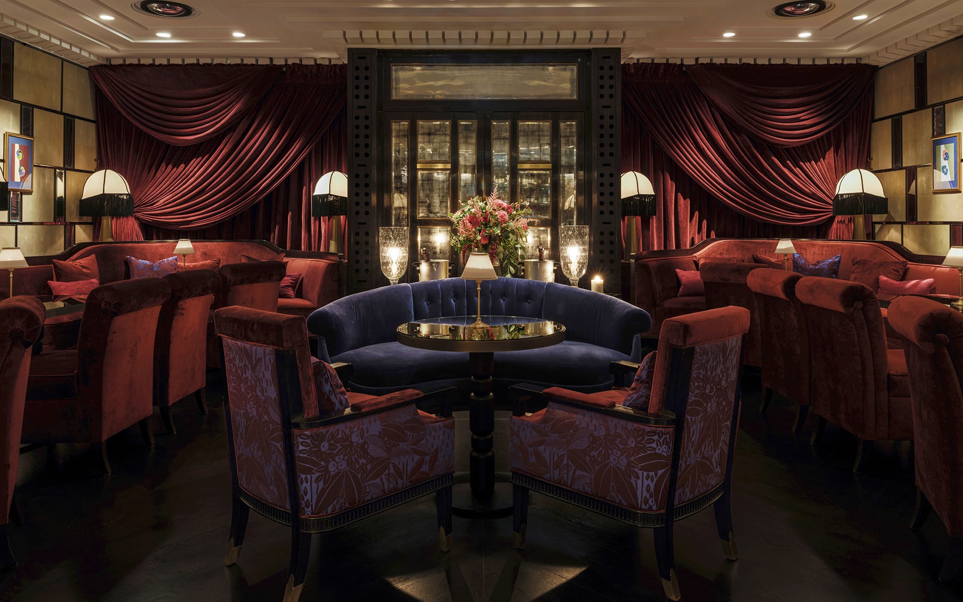 Luxurious lounge with deep red and blue furniture, dim lighting, plush seating, elegant decor, and a floral centerpiece.