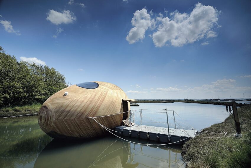 Futuristic wooden pod floating on a river with a small dock, set against a cloudy blue sky and lush greenery.