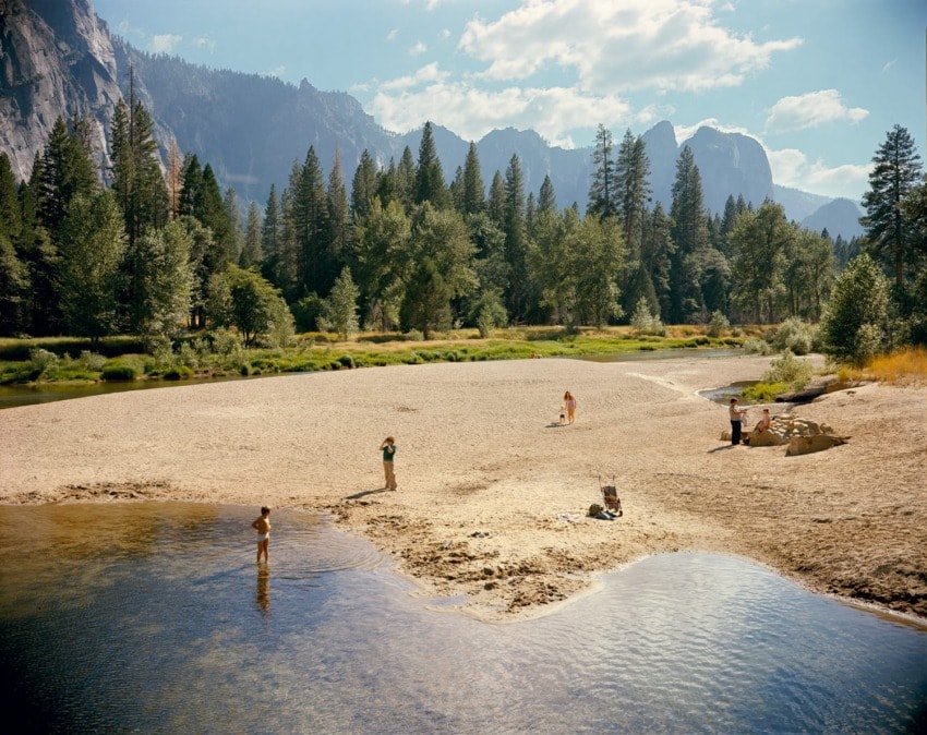 People enjoying a sunny day at a sandy riverside with lush green trees and mountain backdrop in a scenic outdoor setting.