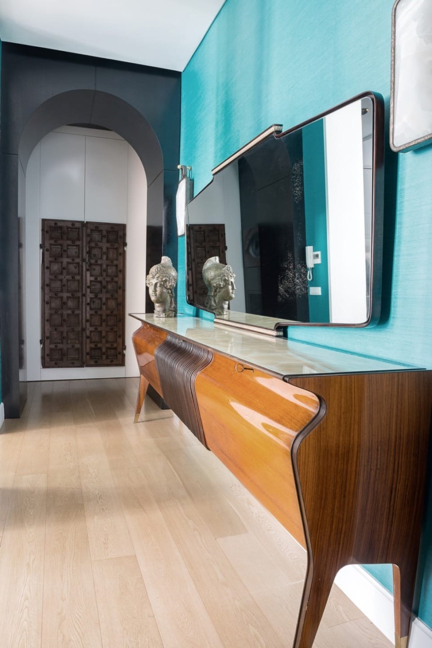 Modern hallway with sleek wooden console, large mirror, teal walls, and decorative elements, leading to a patterned door.