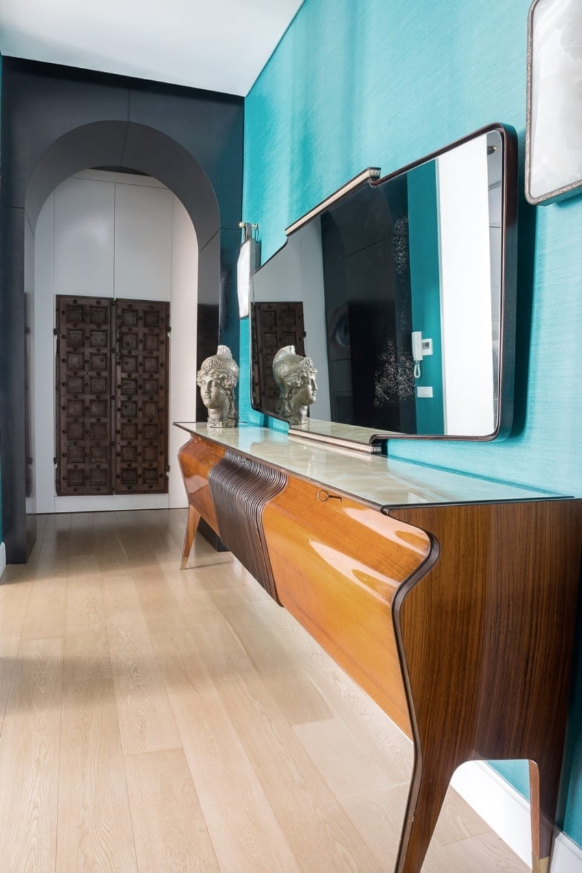 Modern hallway with sleek wooden console, large mirror, teal walls, and decorative elements, leading to a patterned door.