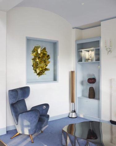 Modern living room with a blue armchair, geometric gold wall art, and light blue shelving.