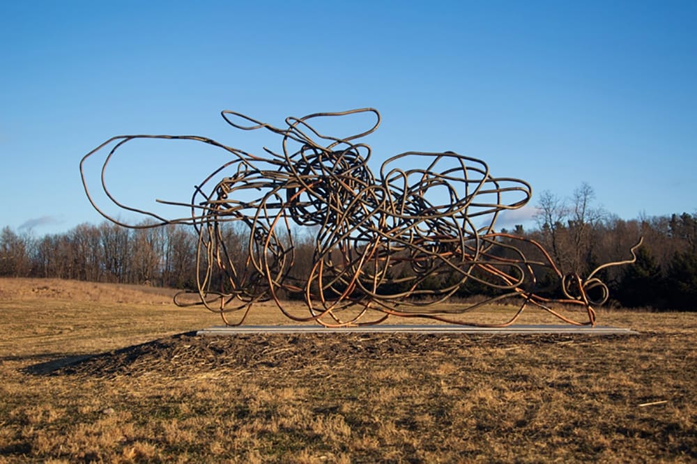Large abstract metal sculpture in a grassy field with trees and a clear blue sky in the background.