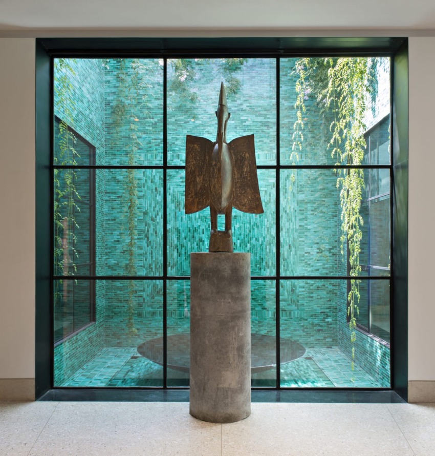 Bronze bird sculpture on stone pedestal in front of large window, with view of teal tiled walls and hanging greenery outside