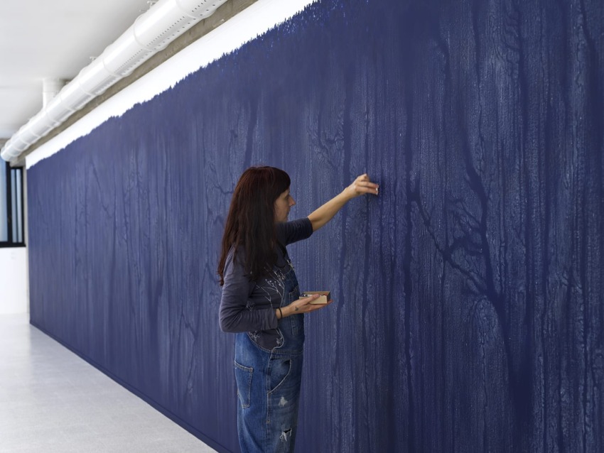 Artist creating a large blue mural on a wall, wearing jeans and a long-sleeve shirt, using a brush for detailed work.